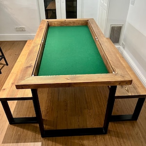 May include: A wooden table with a green felt surface, framed by wood. Two benches are attached, supported by black metal legs. The table is about 183 cm long and 91 cm wide, suitable for games or dining.