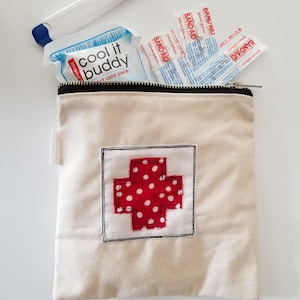 May include: A beige canvas first aid kit with a red cross design with white polka dots. The kit contains Band-Aids, a cold pack, and a white thermometer. The kit has a black zipper.