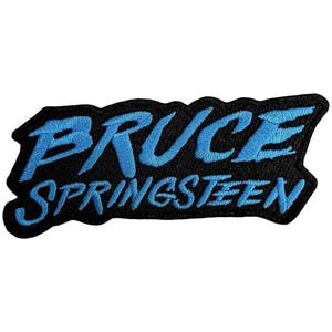May include: A black embroidered patch with the name "BRUCE SPRINGSTEEN" in bright blue, stylized lettering. The patch has a jagged, irregular border.