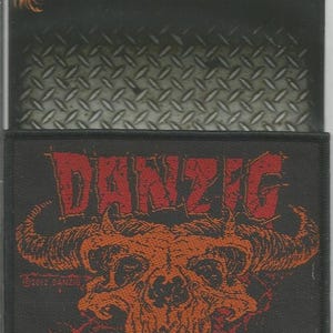 Może przedstawiać: Czarna naszywka do przyszycia z napisem "DANZIG" w czerwonych literach i pomarańczowym wzorem czaszki. Naszywka jest licencjonowana i wyprodukowana w Anglii.
