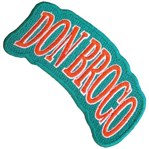 以下が含まれることがあります： 鮮やかなティール色の刺繍パッチ。オレンジ色の筆記体で「DON ROCCO」と書かれ、白い縁取りが施されています。パッチの縁は波型です。