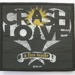 May include: Black square patch with white text reading "CRASH LOVE" and "a fire inside" on a banner. A yellow and white sunburst design is above the letter "A". The patch has a black border and the text "©2010 AFI".