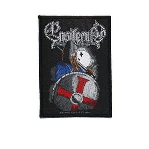 Puede incluir: Parche negro bordado con el nombre de la banda "Ensiferum" en letras góticas blancas. El parche representa a un guerrero con un escudo y un casco. El escudo es gris con una cruz roja. El parche tiene un borde negro y el texto "©2010 ENSIFERUM".