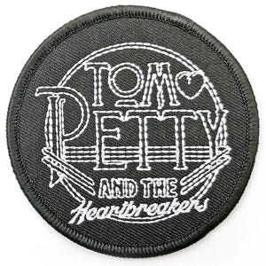 May include: A round, black patch with white embroidered text. The text reads "TOM PETTY" above "AND THE Heartbreakers". The design includes a heart and stylized musical elements. The patch has a stitched border.