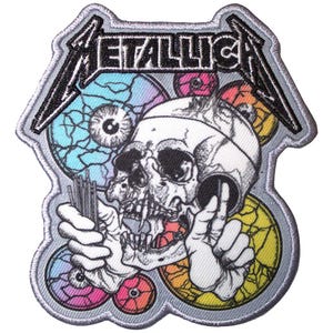 May include: An embroidered patch featuring the word "Metallica" in a stylised font. The patch depicts a skull with a single eye, surrounded by colourful, eye-shaped designs.