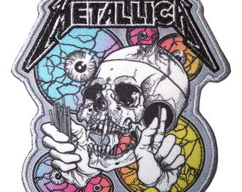 May include: An embroidered patch featuring the Metallica logo in black thread above a white skull illustration. The skull is held by two hands and surrounded by colorful, cracked circular patterns in shades of blue, pink, and yellow.