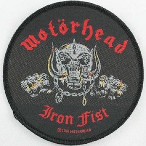 May include: Round black patch featuring the band name "Motörhead" in red, arched above a skull with horns and a chain. Below the skull is "Iron Fist" in red script. The patch includes the copyright notice "©2010 Motörhead".