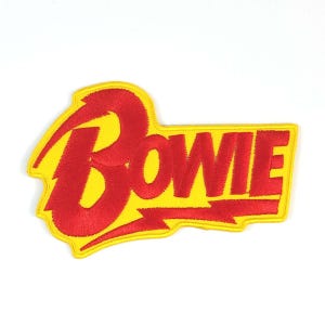 Könnte beinhalten: Gestickter Aufnäher mit dem Namen "BOWIE" in Rot, mit Blitzmotiv. Der Aufnäher hat einen gelben Hintergrund und einen roten Rand. Der Text ist fett und ohne Serifen. Für Kleidung oder Accessoires.