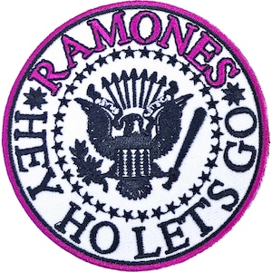 May include: Circular patch featuring the word "RAMONES" in pink, surrounding a black emblem with a baseball bat. The patch has a white background with a pink border and the phrases "HEY HO LET'S GO" in pink.