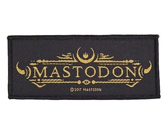 Puede incluir: Parche rectangular negro con bordado dorado. La palabra "MASTODON" destaca, con elementos decorativos arriba y abajo. El texto "© 2017 MASTODON" se encuentra en la parte inferior.