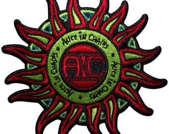 Könnte beinhalten: Gestickter Aufnäher mit einem stilisierten Sonnendesign in Rot, Grün und Schwarz. Der Mittelkreis zeigt die Buchstaben "AIC" in Rot, umgeben vom Text "Alice in Chains" in Grün. Die Sonnenstrahlen sind rot mit grünen und blauen Akzenten.