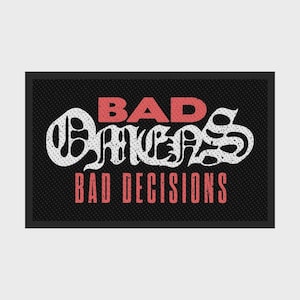 Puede incluir: Felpudo rectangular negro con las palabras "BAD OMENS BAD DECISIONS" en letras rojas y blancas. El texto está estilizado con una fuente única. El fondo es negro texturizado.