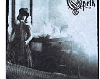 May include: A black and white patch featuring the band name "Opeth" in a stylized font. The patch has a vintage photograph of a person in a dress standing near a table. The image is framed by a black border.