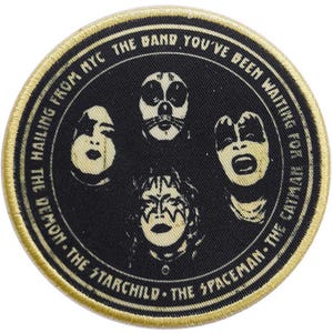 May include: A round, gold-coloured patch with black and white images of four band members. The text around the edge reads "Hailing from NYC the band you've been waiting for... The Demon - The Starchild - The Spaceman - The Catman."