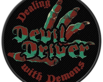 Peut inclure: Écusson rond noir avec les mots "Dealing with Demons" et "Devil Driver" en lettres rouges et turquoise. Une main avec des doigts rouges et turquoise sort du centre. L'écusson a une bordure noire.