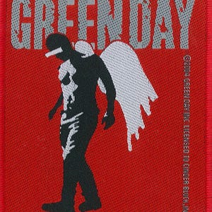 Puede incluir: Un parche tejido rojo con el nombre de la banda "GREEN DAY" en texto blanco. Una silueta negra de una persona con alas de ángel está en el centro del parche.