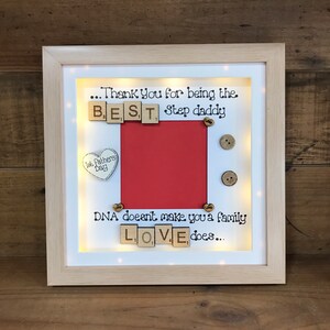 Pu&ograve; includere: Un'immagine incorniciata con uno sfondo rosso e un bordo bianco. La cornice ha una luce all'interno e le parole "...Thank You for being the BEST Step daddy" sono scritte con tessere di lettere in legno. Le parole "DNA doesn't make you a family LOVE does..." sono anche scritte con tessere di lettere in legno.