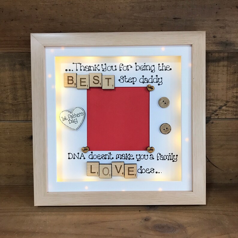Pu&ograve; includere: Un'immagine incorniciata con uno sfondo rosso e un bordo bianco. La cornice ha una luce all'interno e le parole "...Thank You for being the BEST Step daddy" sono scritte con tessere di lettere in legno. Le parole "DNA doesn't make you a family LOVE does..." sono anche scritte con tessere di lettere in legno.