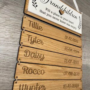 May include: Wooden sign with the text "Grandchildren Fill a place in your heart that you never knew was empty."  The sign has five wooden slats with names and dates engraved on each slat. The names are Tillie, Tyler, Daisy, Rocco, and Wynter. The dates are 31/12/2013, 15/08/2014, 12/07/2015, 07/09/2018, and 10/10/2023.