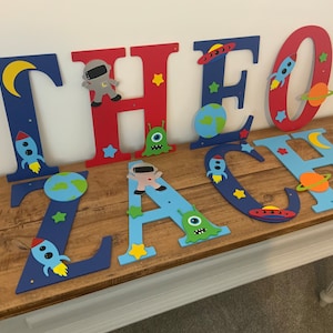 May include: Blue, red, and light blue wooden letters spelling the name "THEO ZACH" decorated with space-themed cutouts, including rockets, planets, stars, astronauts, and aliens.