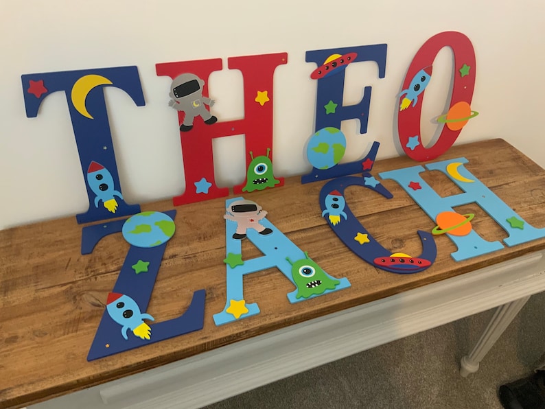 May include: Blue, red, and light blue wooden letters spelling the name "THEO ZACH" decorated with space-themed cutouts, including rockets, planets, stars, astronauts, and aliens.