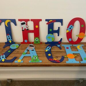 May include: Wooden letters spelling "THEO ZACH" in blue, red, and white paint. Each letter is decorated with space-themed cutouts, including rockets, planets, stars, and astronauts.