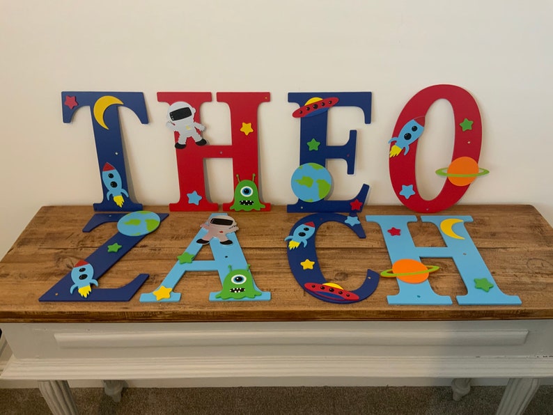 May include: Wooden letters spelling "THEO ZACH" in blue, red, and white paint. Each letter is decorated with space-themed cutouts, including rockets, planets, stars, and astronauts.