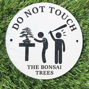 May include: A white, circular sign with black text and illustrations. The text reads "DO NOT TOUCH THE BONSAI TREES." The illustration depicts a bonsai tree, a person reaching for it, and another person with a baseball bat.