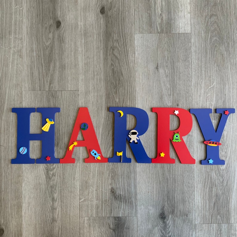 May include: Red and blue wooden letters spelling the name "HARRY" with space-themed decorations. The letters are decorated with stars, planets, rockets, and astronauts.
