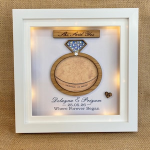 May include: A white shadow box frame containing a wooden ring-shaped cutout with a map inlay, a diamond-shaped piece with blue crystals, and a wooden plaque reading She Said Yes. Text reads Delayna & Priyam, 25.05.26, and Where Forever Began.