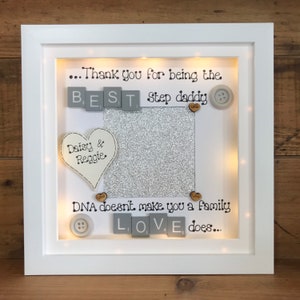 Pu&ograve; includere: Una cornice bianca con uno sfondo di glitter argentati e un messaggio che recita "...Grazie per essere il MIGLIOR pap&agrave;. Il DNA non fa una famiglia, l'AMORE s&igrave;..." La cornice ha un cuore con le parole "Daisy & Reggie" e bottoni con la parola "Amore" su di essi.