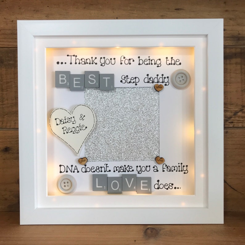 Pu&ograve; includere: Una cornice bianca con uno sfondo di glitter argentati e un messaggio che recita "...Grazie per essere il MIGLIOR pap&agrave;. Il DNA non fa una famiglia, l'AMORE s&igrave;..." La cornice ha un cuore con le parole "Daisy & Reggie" e bottoni con la parola "Amore" su di essi.