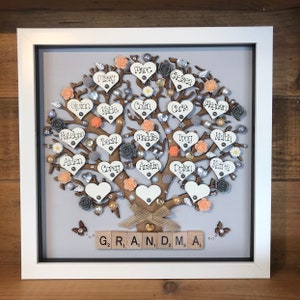 May include: A framed wooden family tree with names written on heart-shaped pieces of wood. The tree is decorated with peach and gray flowers, pearls, and rhinestones. The base of the tree has the word "GRANDMA" spelled out with wooden letter tiles.