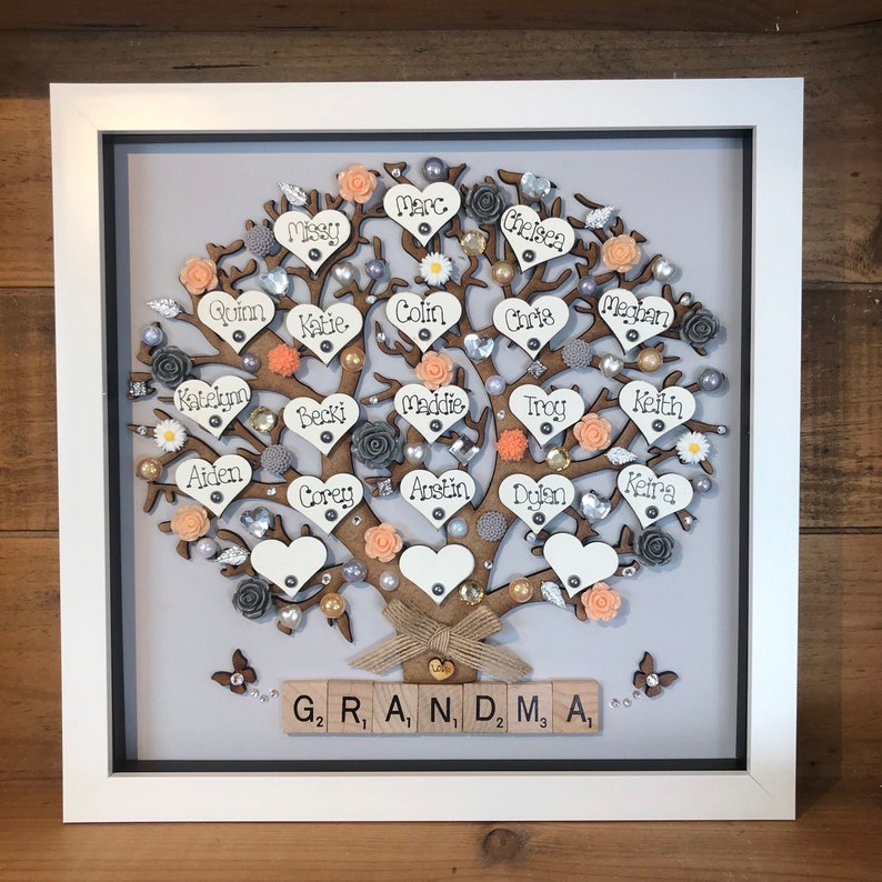 May include: A framed wooden family tree with names written on heart-shaped pieces of wood. The tree is decorated with peach and gray flowers, pearls, and rhinestones. The base of the tree has the word "GRANDMA" spelled out with wooden letter tiles.