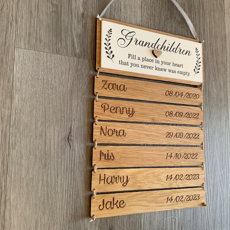 May include: Wooden sign with the text "Grandchildren Fill a place in your heart that you never knew was empty."  The sign has a small heart and a list of names and dates. The names are Zara, Penny, Nora, Iris, Harry, and Jake.