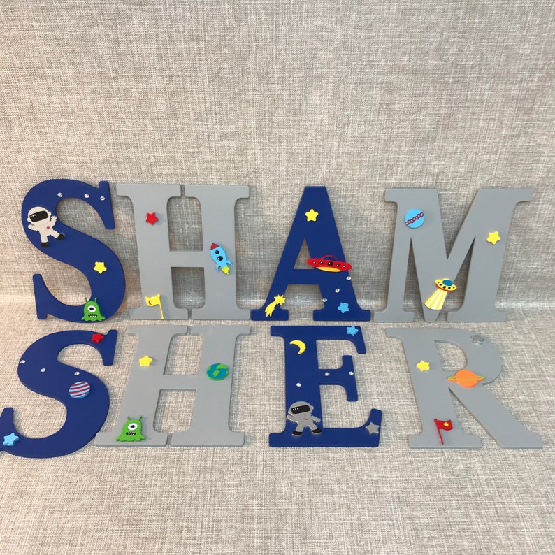 May include: A set of wooden letters spelling out the name "SHAMER" in blue and gray paint. The letters are decorated with space-themed embellishments, including stars, planets, rockets, and astronauts.