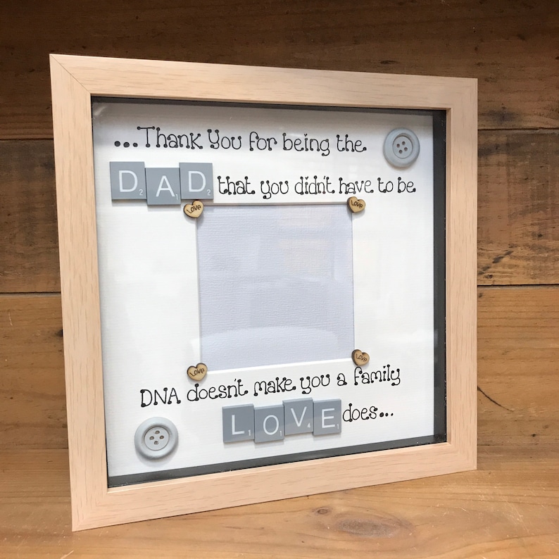 Pu&ograve; includere: Una cornice per foto bianca con una cornice in legno chiaro. La cornice ha uno sfondo bianco con il testo "...Thank You for being the. DAD. that you didn't have to be Love DNA doesn't make you a family LOVE does..." La cornice ha un piccolo spazio per foto al centro.