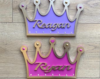 May include: Two wooden princess crowns with gold trim and pink and purple paint. The top crown says "Reagan" and the bottom crown says "Ryan".
