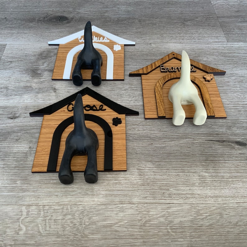 May include: Three wooden dog house shaped coat hooks with black, white, and brown paint. The hooks are shaped like dog tails and are labeled "Wibbles", "Bramble", and "Goose".