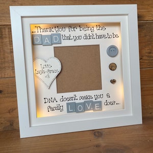 Pu&ograve; includere: Cornice per foto bianca con un passepartout marrone chiaro e un'apertura per foto. La cornice ha uno sfondo bianco con il testo "...Thank You for being the DAD, that you didn't have to be" e "DNA doesn't make you a family LOVE does..." in nero. La cornice ha un cuore con il testo "Love Layla-Grace <3" e alcuni bottoni.