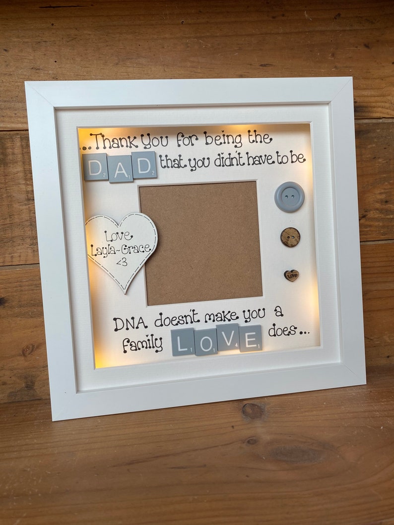 Pu&ograve; includere: Cornice per foto bianca con un passepartout marrone chiaro e un'apertura per foto. La cornice ha uno sfondo bianco con il testo "...Thank You for being the DAD, that you didn't have to be" e "DNA doesn't make you a family LOVE does..." in nero. La cornice ha un cuore con il testo "Love Layla-Grace <3" e alcuni bottoni.