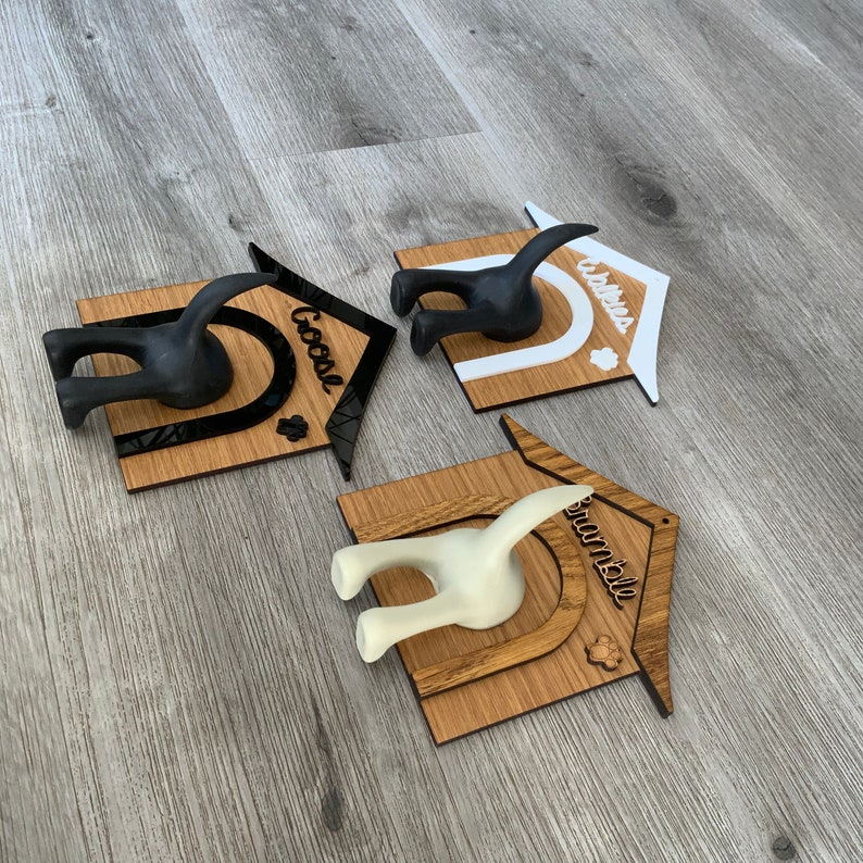 May include: Three wooden dog house shaped coat hooks with the names "Goose", "Walter", and "Snowball" written on them. Each hook has a black or white dog tail sticking out of the back.