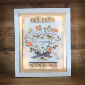 May include: A white frame with a gold border, containing a wooden tree with gold and peach embellishments. The tree has two hearts with the names "George" and "Margaret" and the date "5th September 2020". The words "GOLDEN WEDDING" are spelled out with wooden tiles.