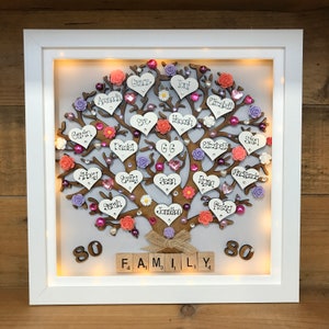 May include: A white framed illuminated wooden family tree with pink, purple, and orange hearts and flowers. The names of family members are written on the hearts. The tree is decorated with small white flowers and rhinestones. The bottom of the frame has the word "FAMILY" spelled out with wooden letter tiles and the numbers "80" on either side.