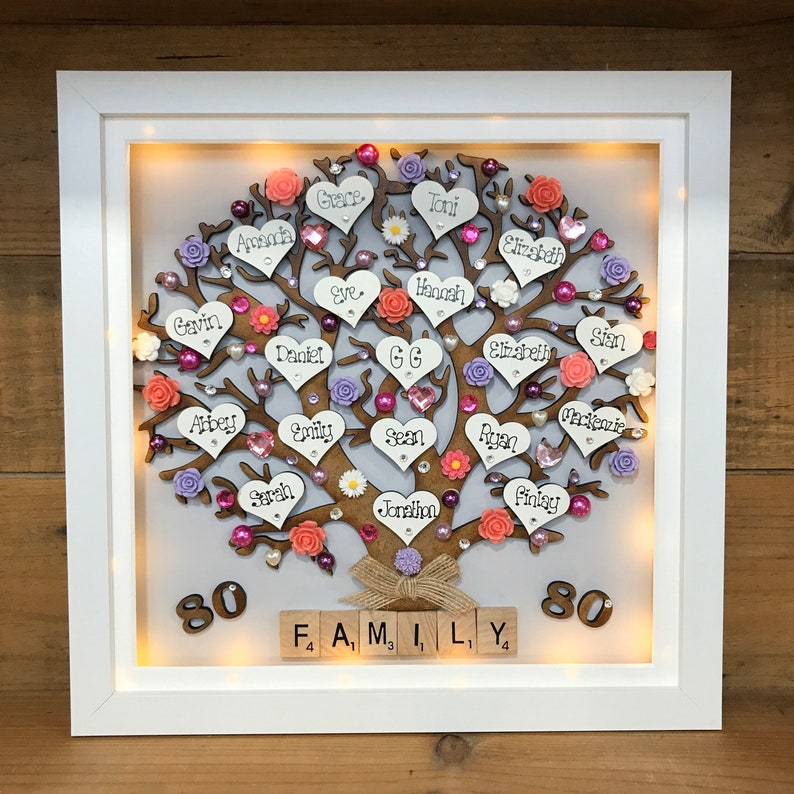 May include: A white framed illuminated wooden family tree with pink, purple, and orange hearts and flowers. The names of family members are written on the hearts. The tree is decorated with small white flowers and rhinestones. The bottom of the frame has the word "FAMILY" spelled out with wooden letter tiles and the numbers "80" on either side.
