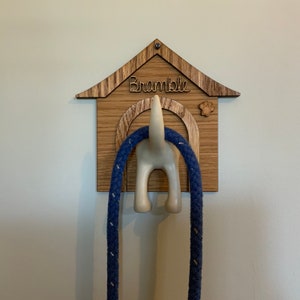 May include: Wooden dog house shaped wall hook with a white dog tail sticking out the back. The hook is labeled "Bramble" and has a paw print design. A blue rope leash is hanging from the hook.