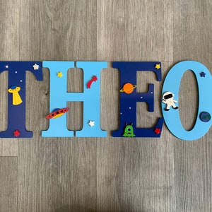 May include: Blue and light blue wooden letters spelling the name "THEO" with space-themed decorations, including stars, planets, a rocket, an alien, and an astronaut.