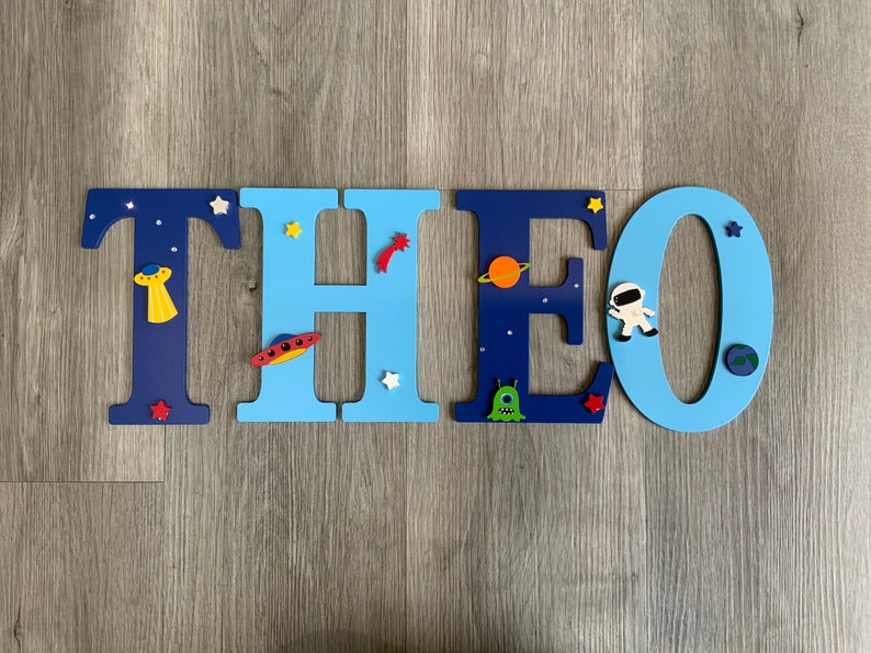 May include: Blue and light blue wooden letters spelling the name "THEO" with space-themed decorations, including stars, planets, a rocket, an alien, and an astronaut.