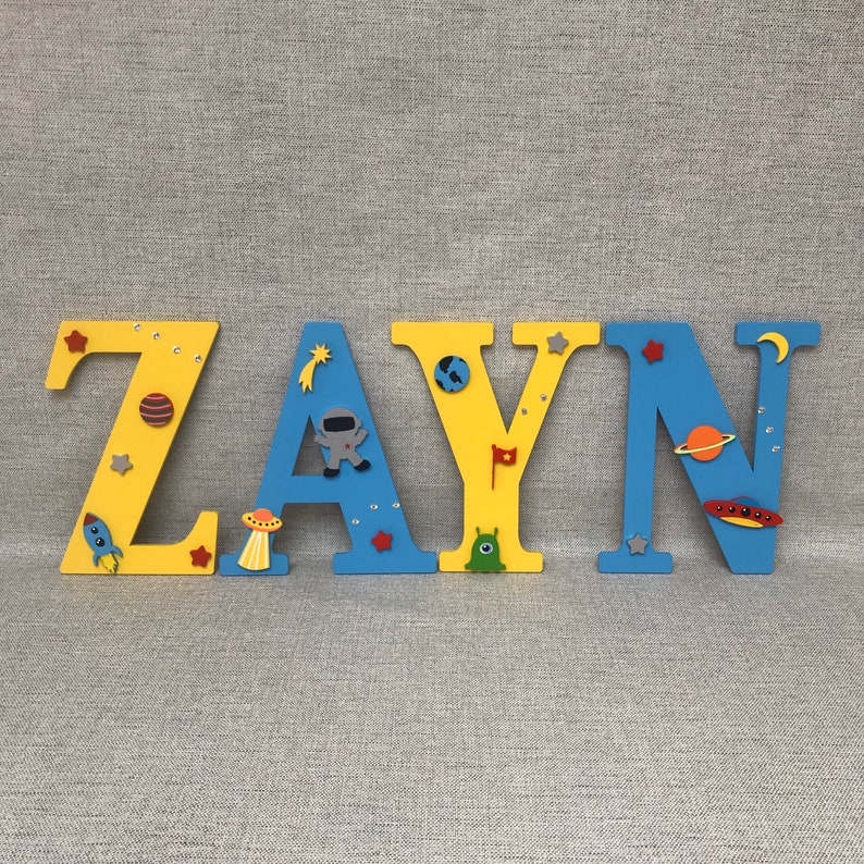 May include: Wooden letters spelling the name "ZAYN" in yellow and blue paint. Each letter is decorated with space-themed embellishments, including stars, planets, rockets, and aliens.
