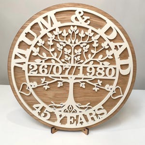 May include: A circular wooden plaque with a white tree design, the text "MUM & DAD" and "26/07/1980" is cut out. The plaque also features the text "45 YEARS" and is displayed on a wooden stand.